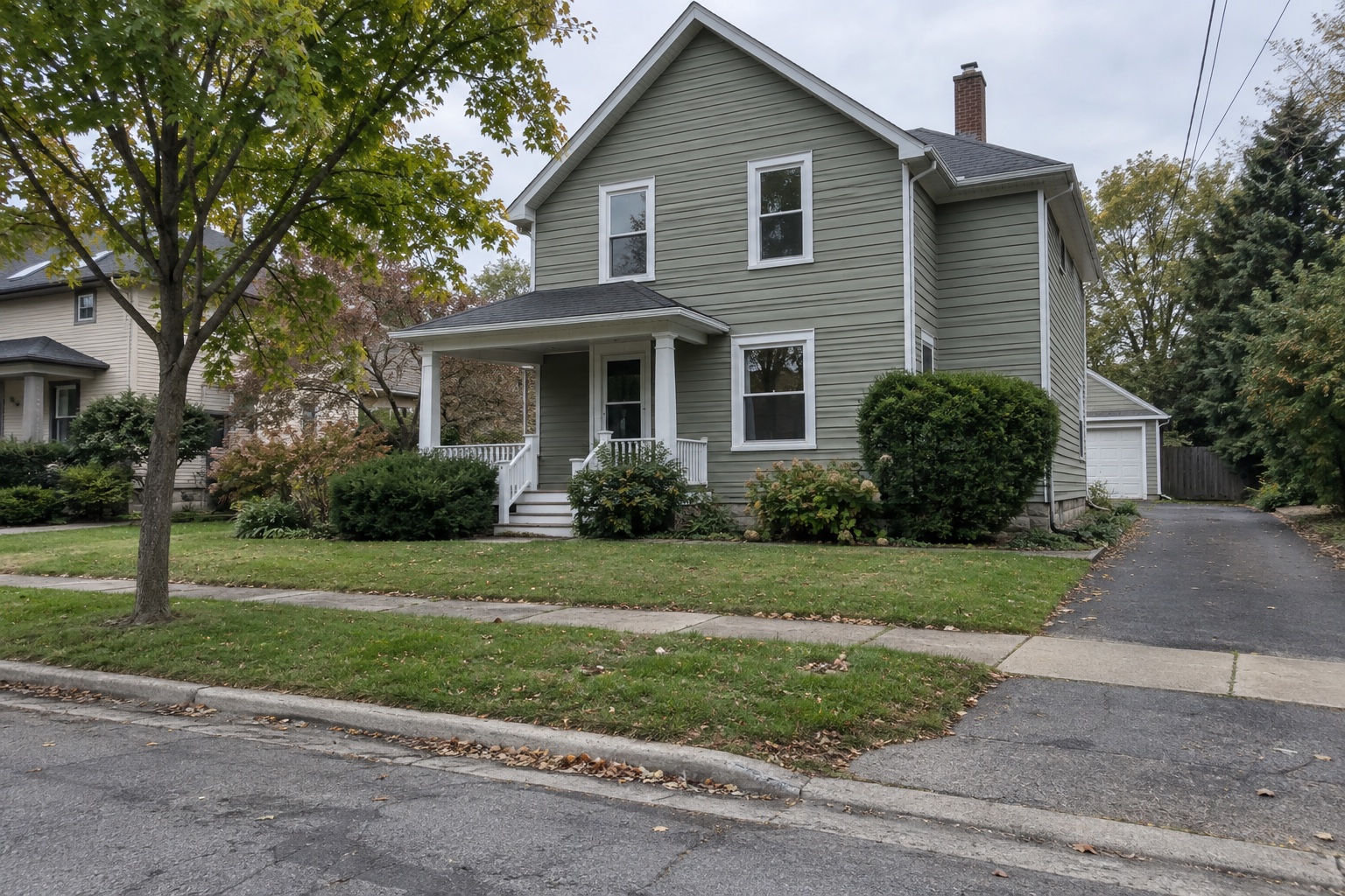 Street-facing view of a sage-gray two-story house with porch, driveway, sidewalk and curb in overcast daylight